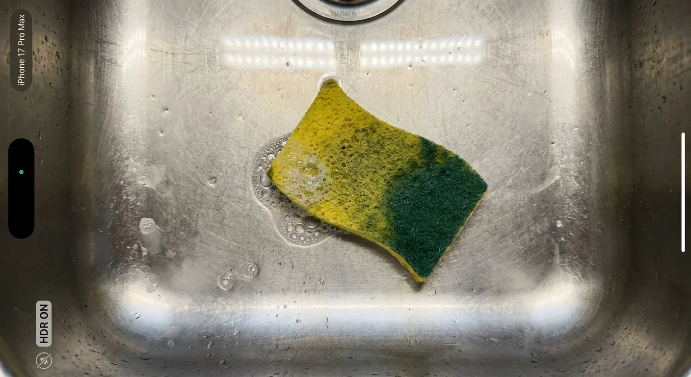 A yellow and green sponge with soap suds sits in a stainless steel kitchen sink.