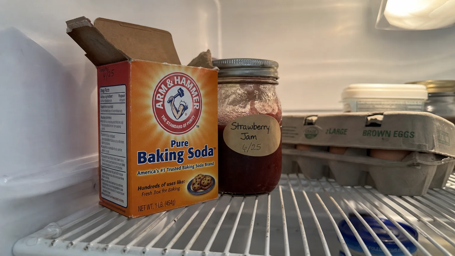 A classic orange cardboard box of baking soda sitting on a wire shelf inside a refrigerator next to a jar of jam.