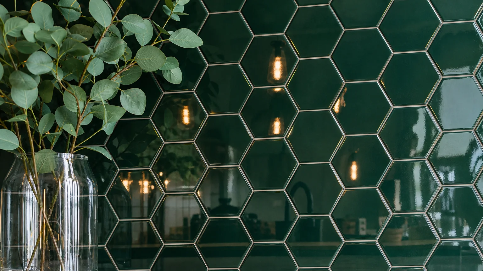 A close-up of glossy, deep forest green hexagon tiles with thin, clean grey grout lines reflecting warm kitchen lighting.