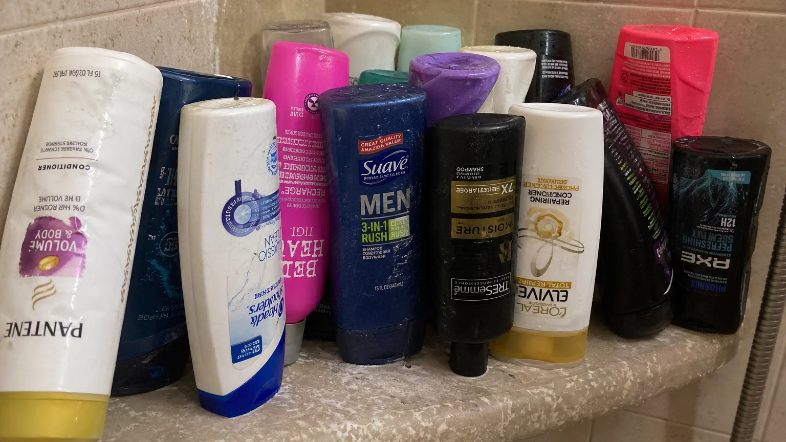 A crowded shower shelf filled with numerous nearly-empty, upside-down plastic bottles.