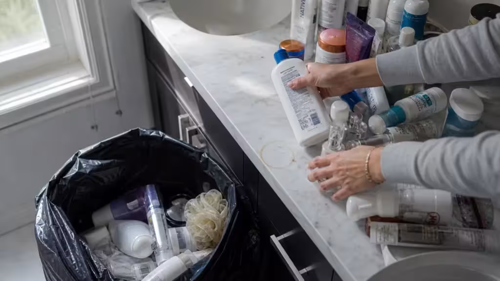 An overhead shot of a person sorting through bathroom clutter on a marble counter, tossing old items into a black garbage bag.