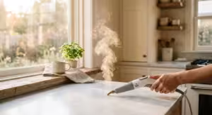 A plume of white steam rises from a steam cleaner nozzle on a sunlit marble kitchen countertop.
