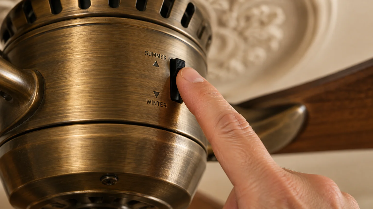 A finger adjusts the black directional switch on an antique brass ceiling fan motor marked Summer and Winter.