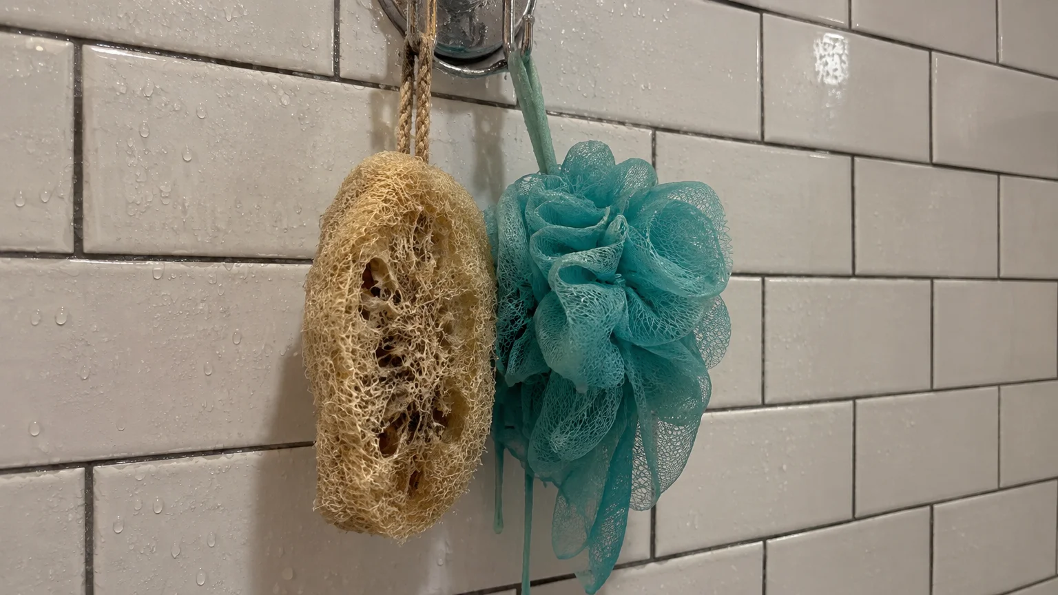 A natural fiber loofah sponge and a turquoise mesh bath pouf hanging from a metal hook against white subway shower tile.