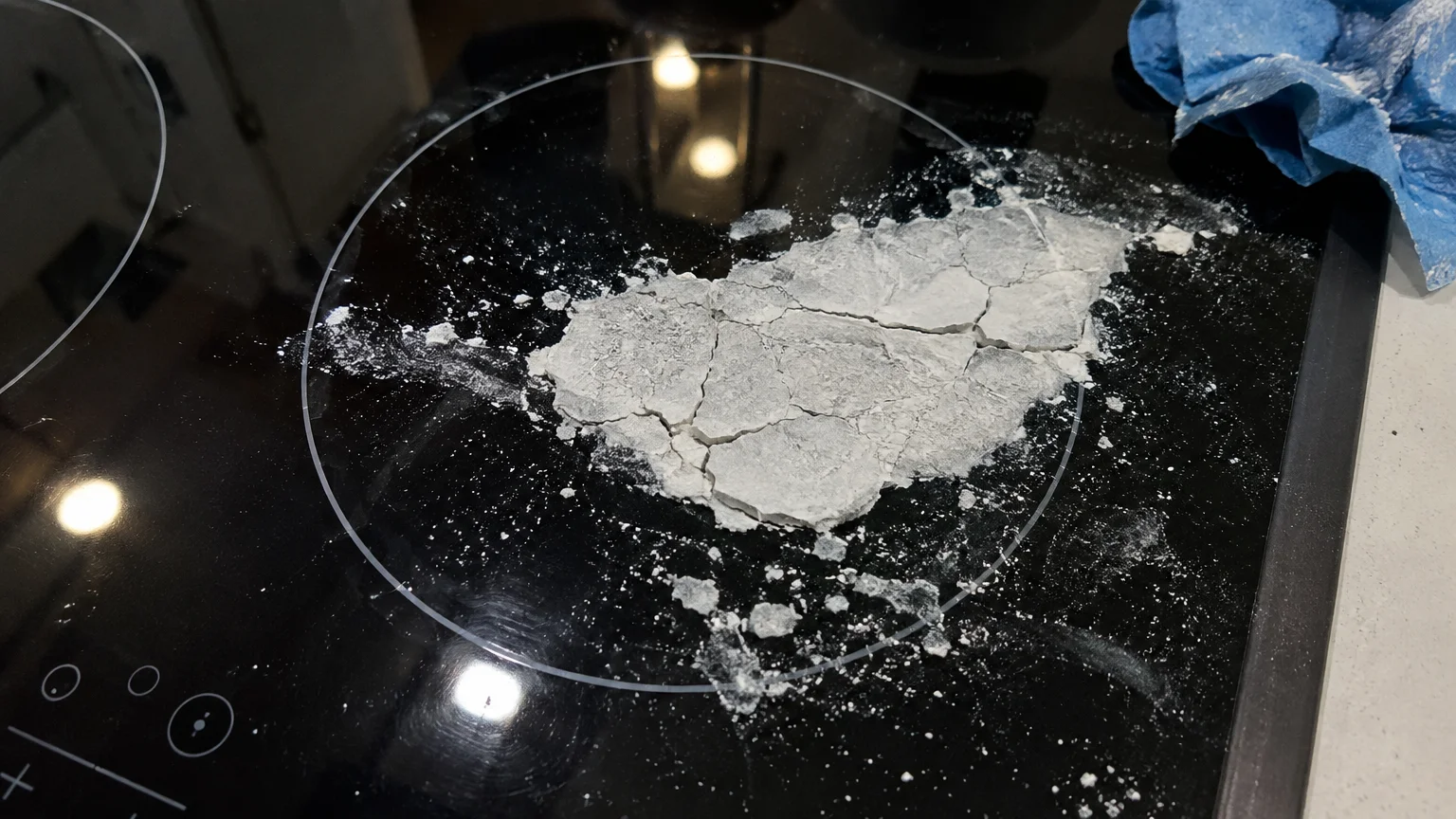 Cracked, dried white baking soda paste caked onto a black glass ceramic stovetop burner beside a blue cloth.