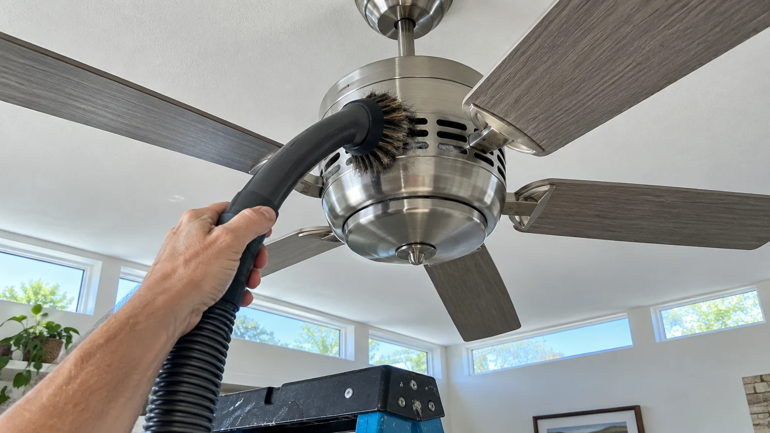 Hand holding a vacuum dusting brush attachment against the vent slots of a brushed metal ceiling fan.