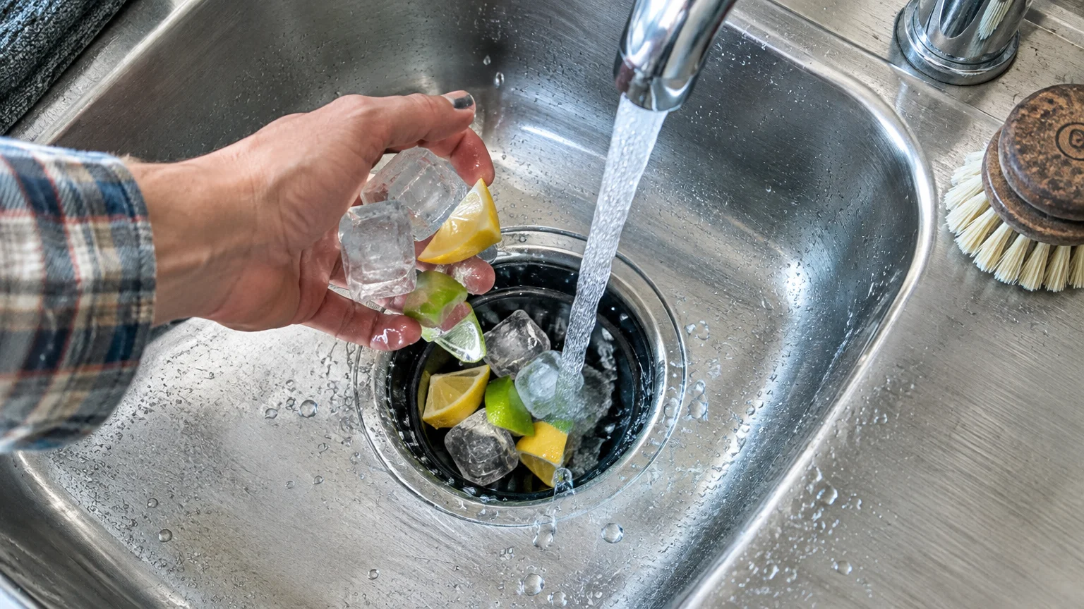 Hand dropping ice cubes, lemon wedges, and lime pieces into a stainless steel sink drain with running water.