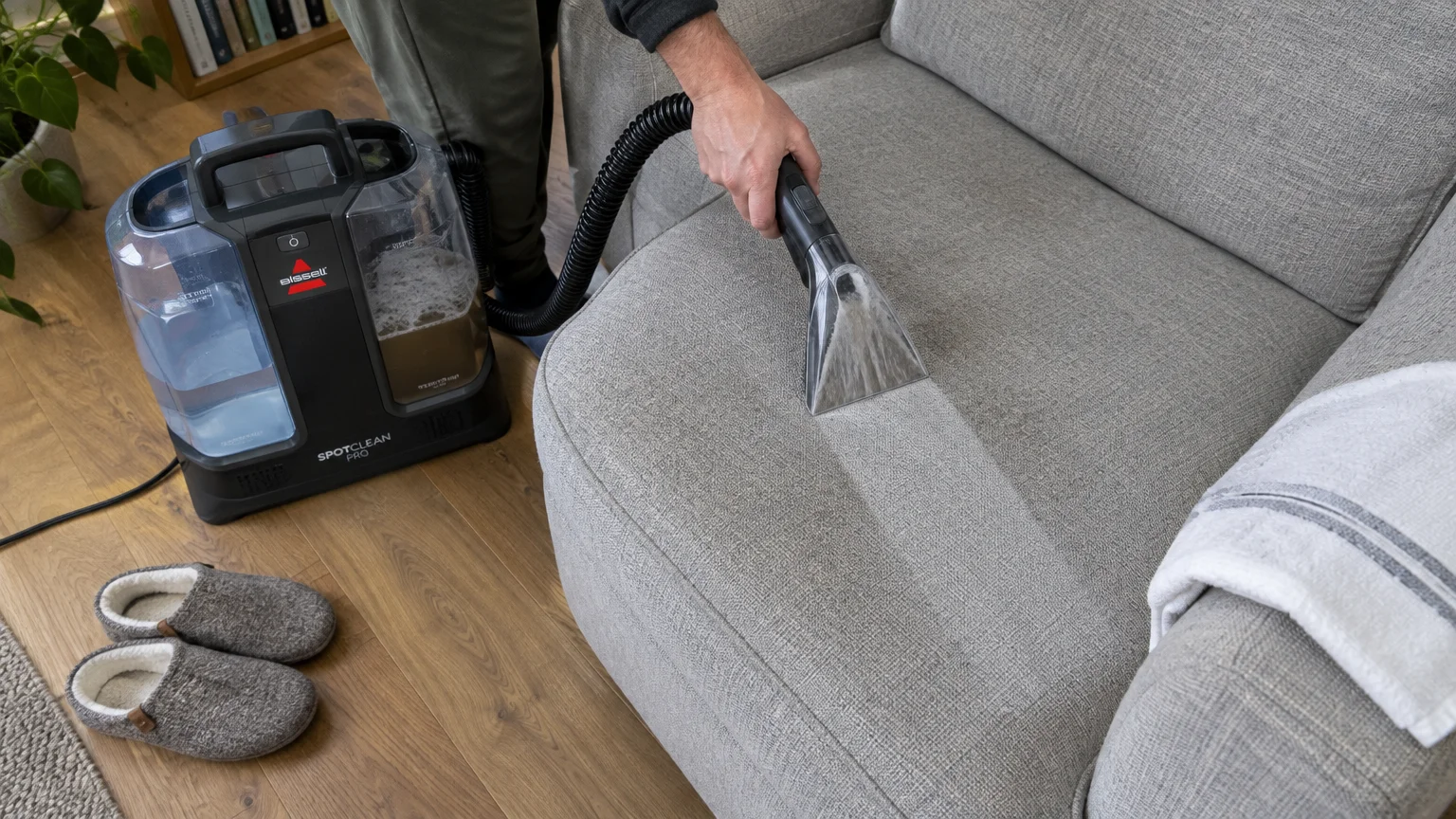 A person using a portable carpet and upholstery extractor vacuum to clean a grey fabric armchair cushion.