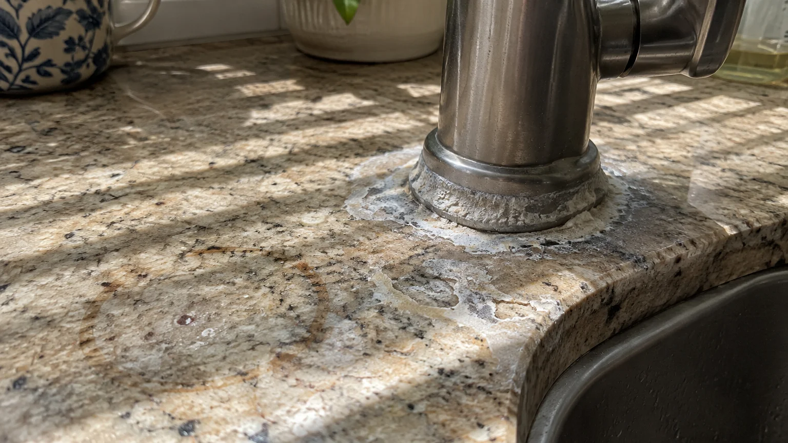 Hard water stains and white mineral crust buildup around a kitchen faucet base on a granite countertop.