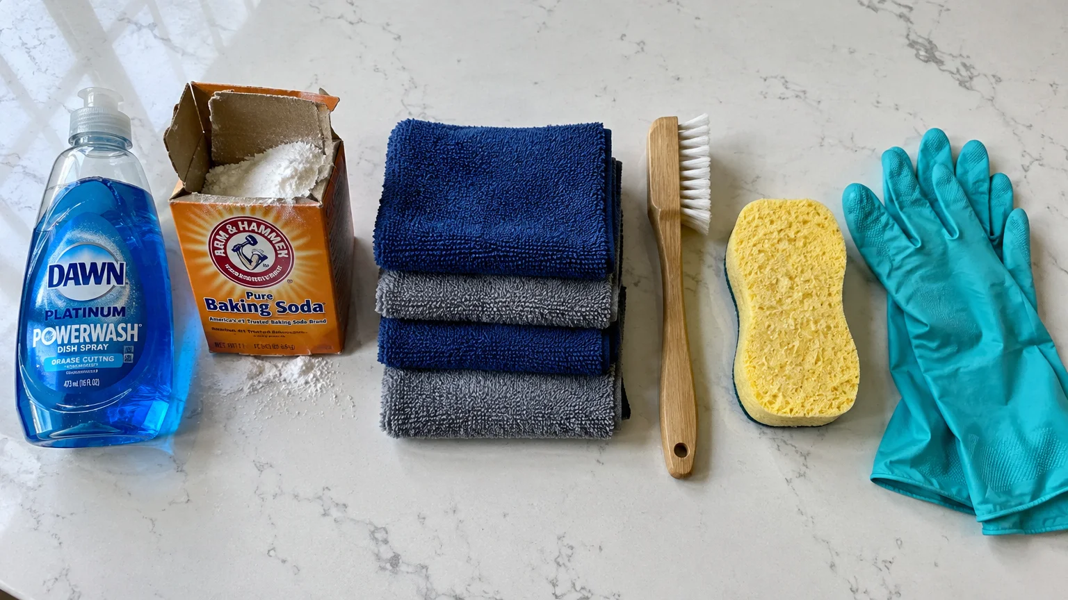 Dish soap, baking soda, folded microfiber towels, a wooden scrub brush, yellow sponge, and rubber gloves on a counter.