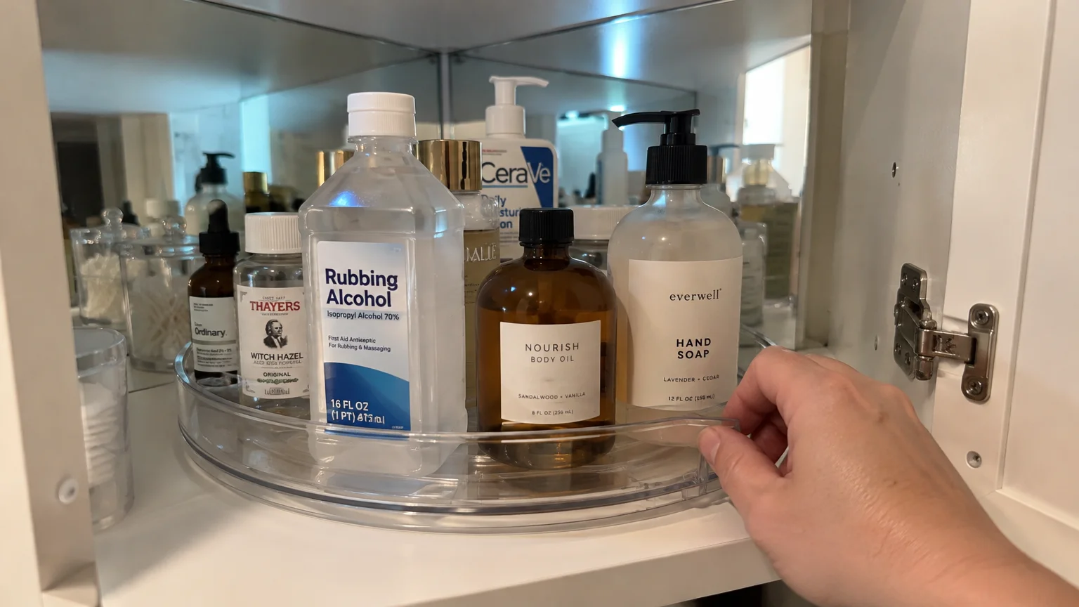 Hand rotating a clear plastic lazy susan filled with bottles inside a mirrored corner medicine cabinet.