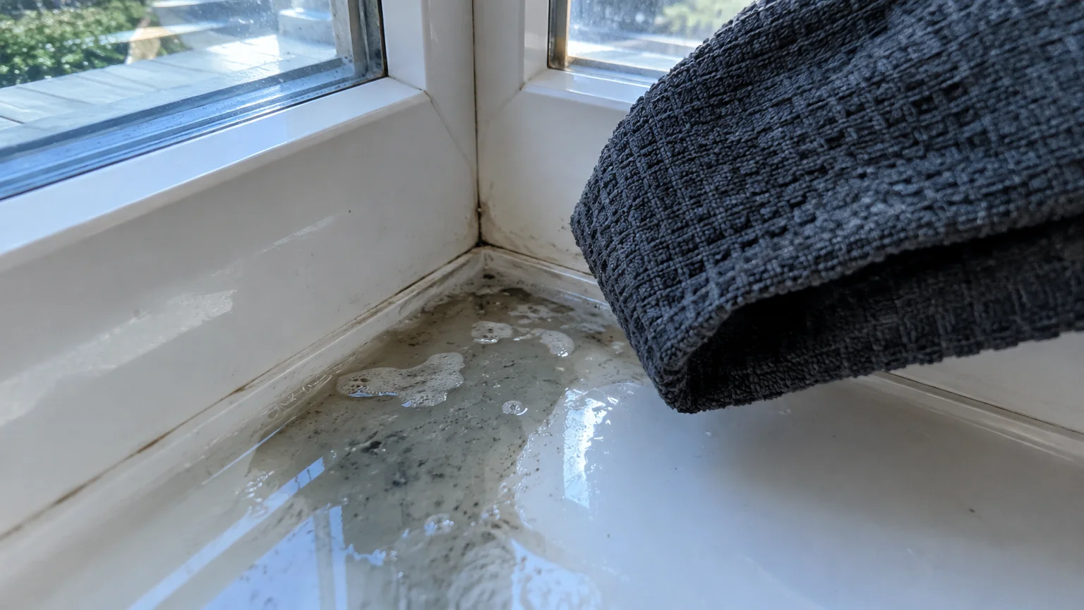 Dirty, soapy rinse water pooling in the corner of a white window frame next to a grey microfiber cloth.