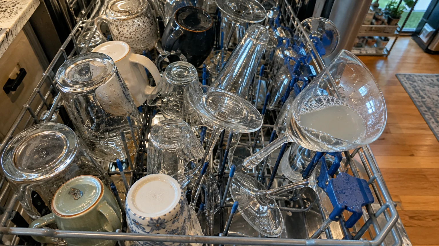 A crowded dishwasher rack filled with mugs, upside-down tumblers, and a wine glass filled with standing cloudy water.