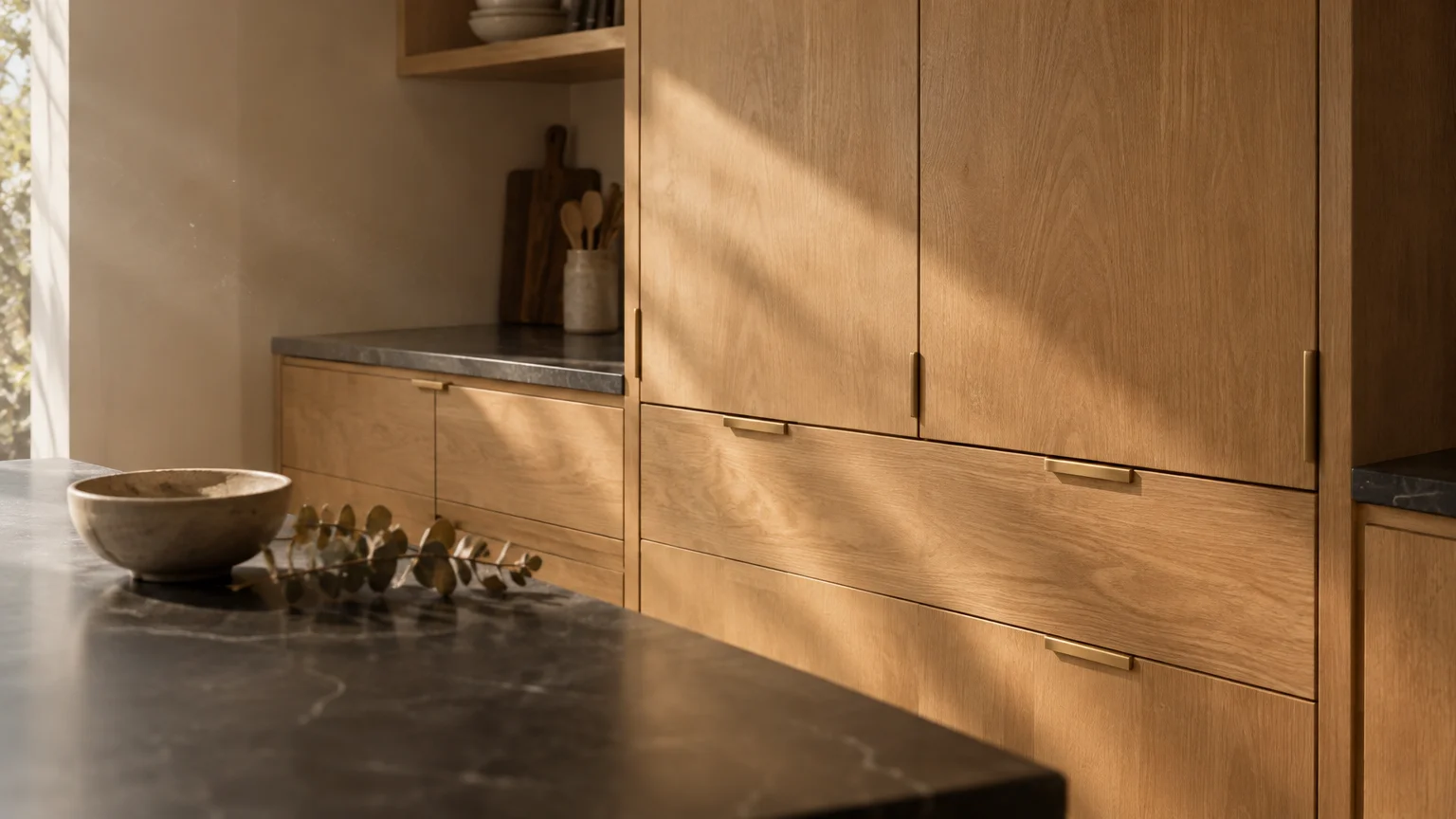 Sunlight illuminates modern light wood kitchen cabinets paired with dark marble countertops.